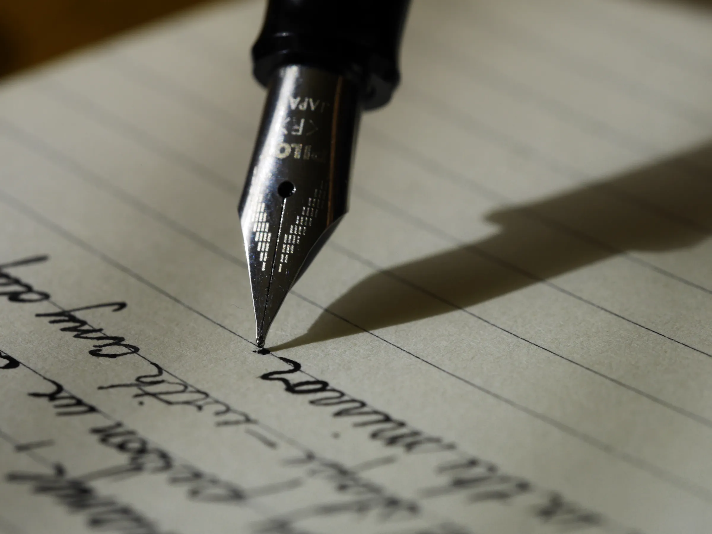 Close-up of a fountain pen nib writing cursive script on lined paper, its shadow falling across the page.