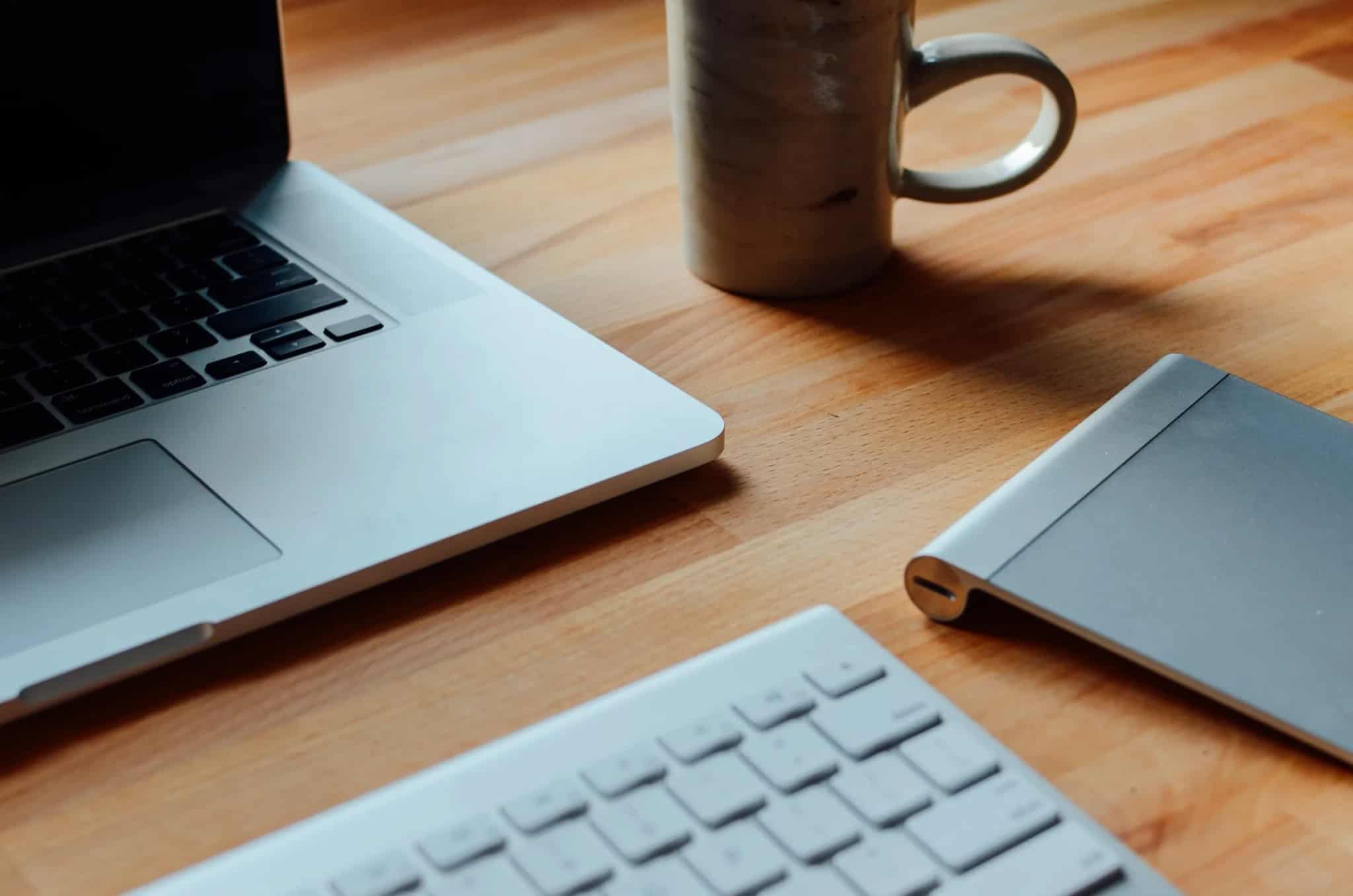 Close-up of a laptop, keyboard, and a mug of coffee on a wooden desk.