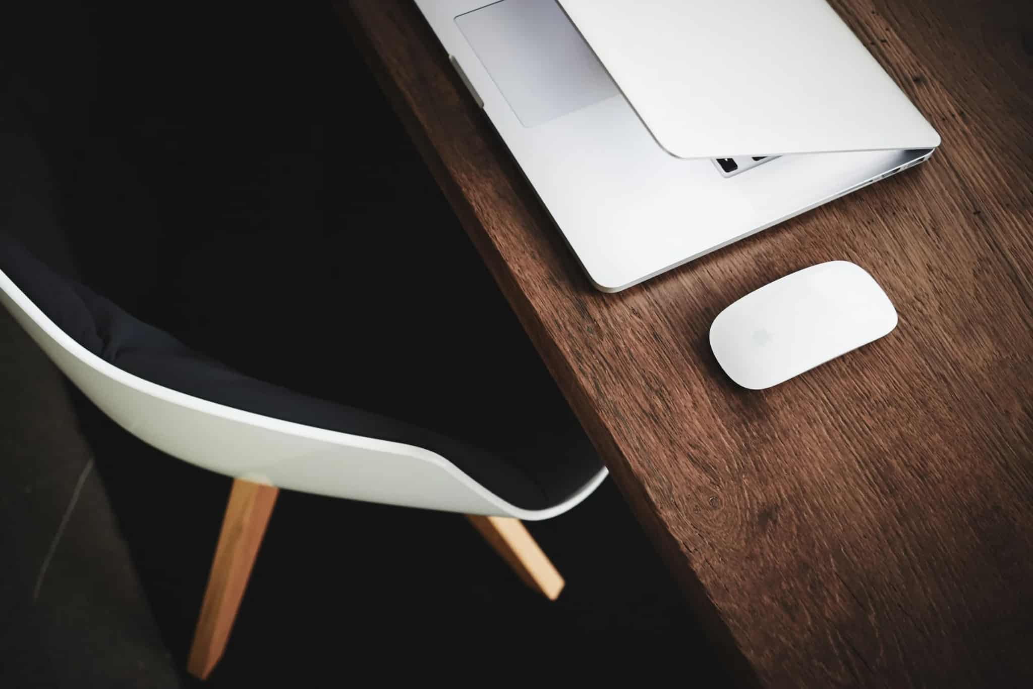 A laptop lid caught mid-close beside an office chair, its screen still glowing with a webpage as a wireless mouse sits nearby on a dark desk.