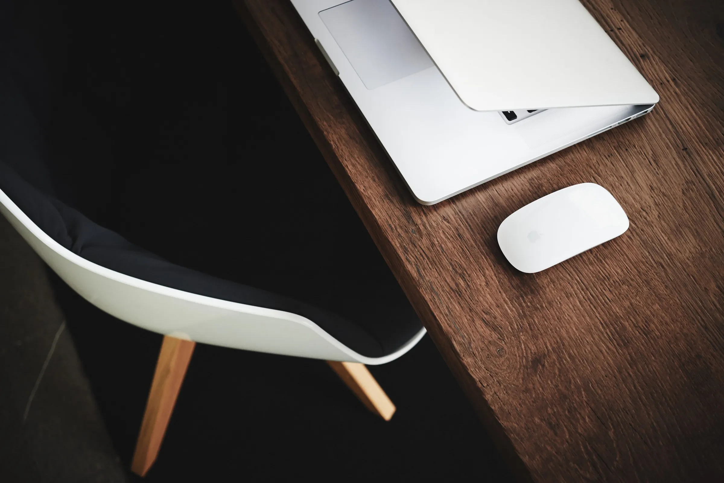 A laptop lid caught mid-close beside an office chair, its screen still glowing with a webpage as a wireless mouse sits nearby on a dark desk.