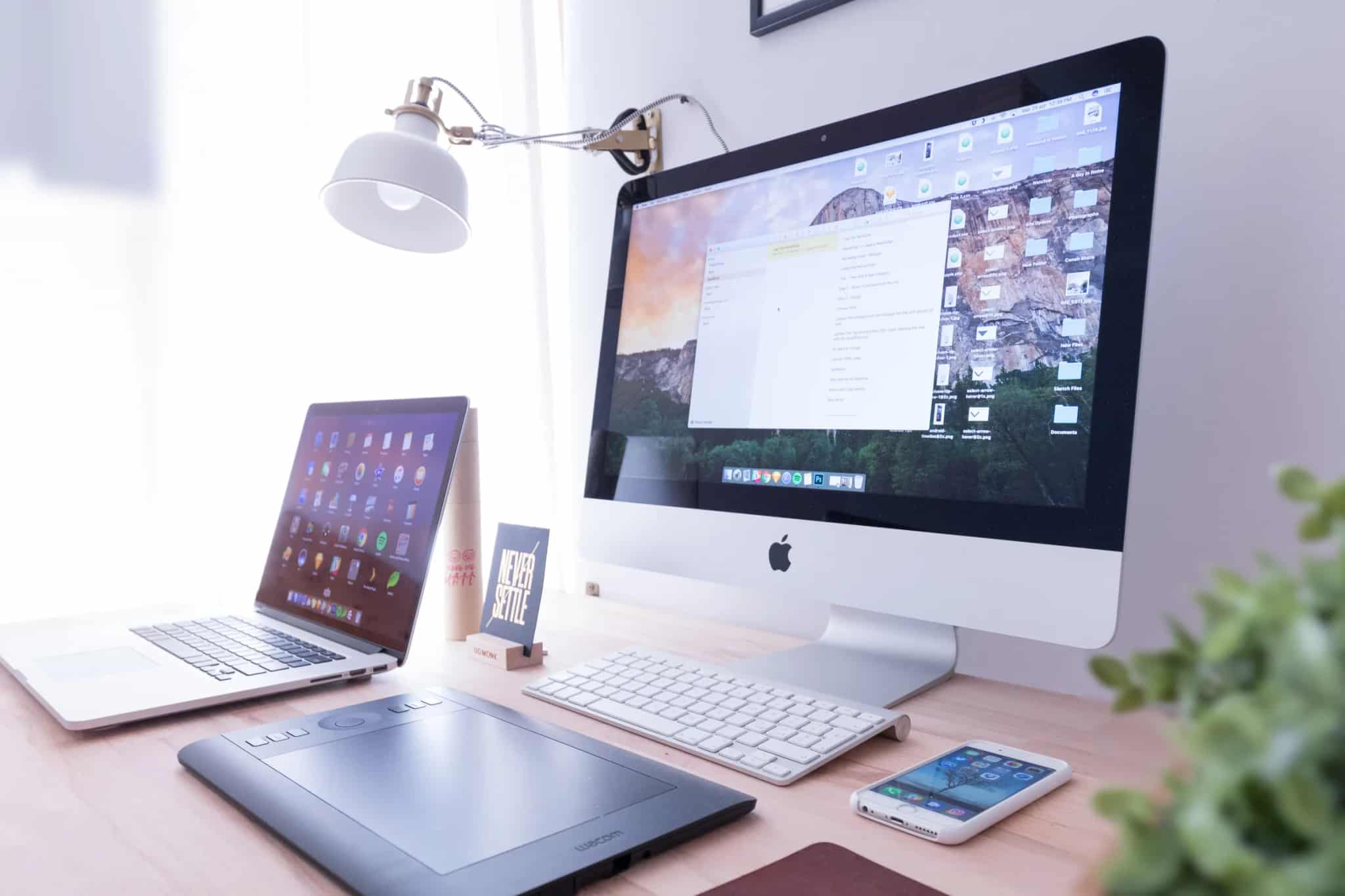 An iMac desktop showing an open document window, a MacBook displaying a grid of colorful app icons, and an iPhone resting nearby on a wooden desk.