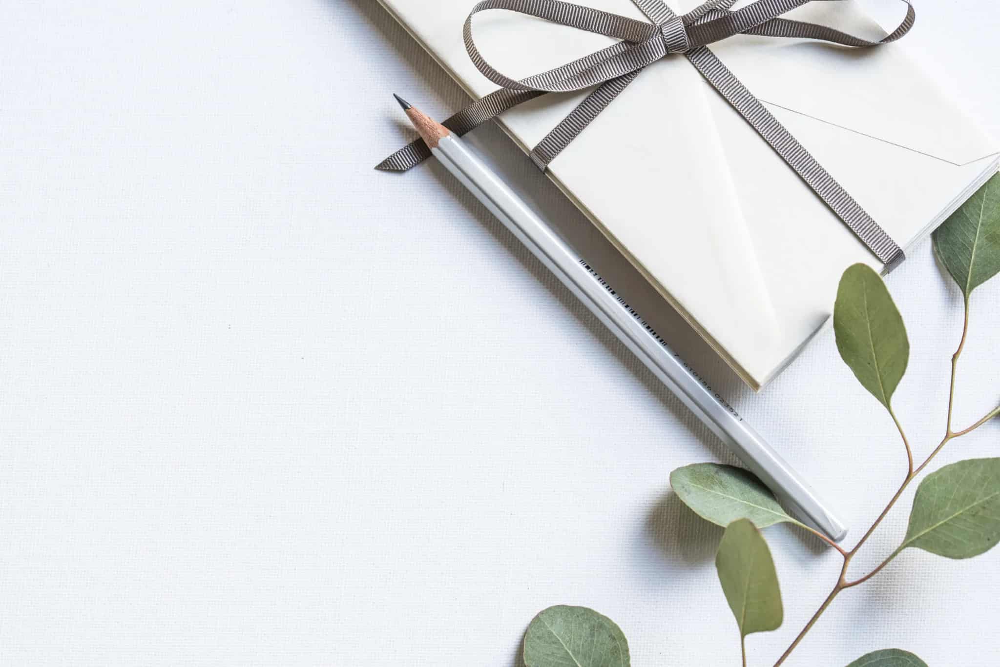 A cream envelope tied with a gray ribbon bow rests beside a silver pencil and a sprig of eucalyptus leaves on a white surface.