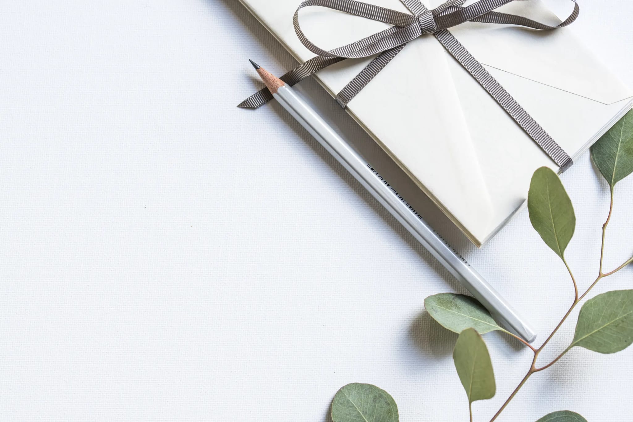 A cream envelope tied with a gray ribbon bow rests beside a silver pencil and a sprig of eucalyptus leaves on a white surface.