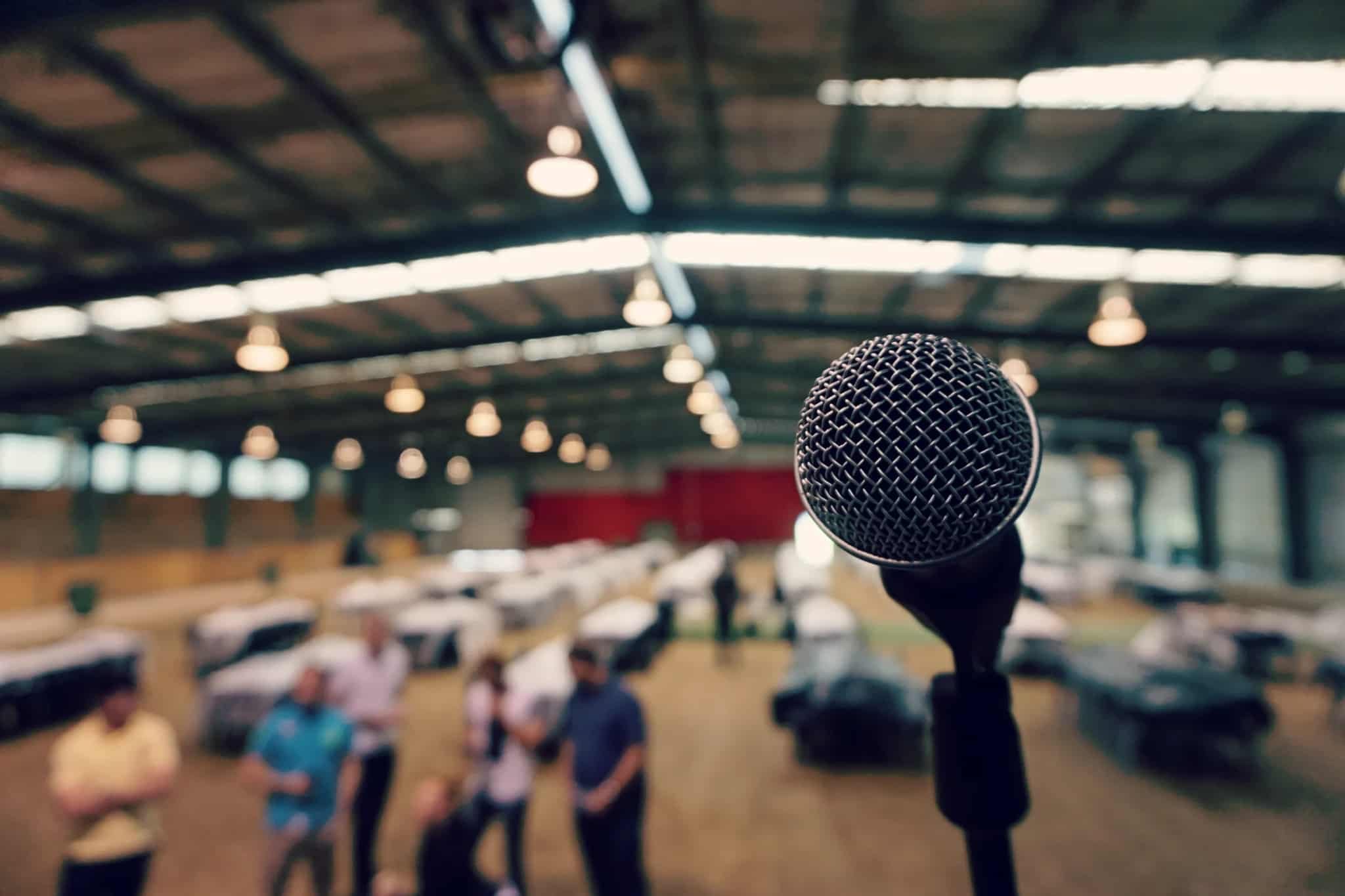 Close-up of a microphone on a stand with blurred people in a large hall behind it.