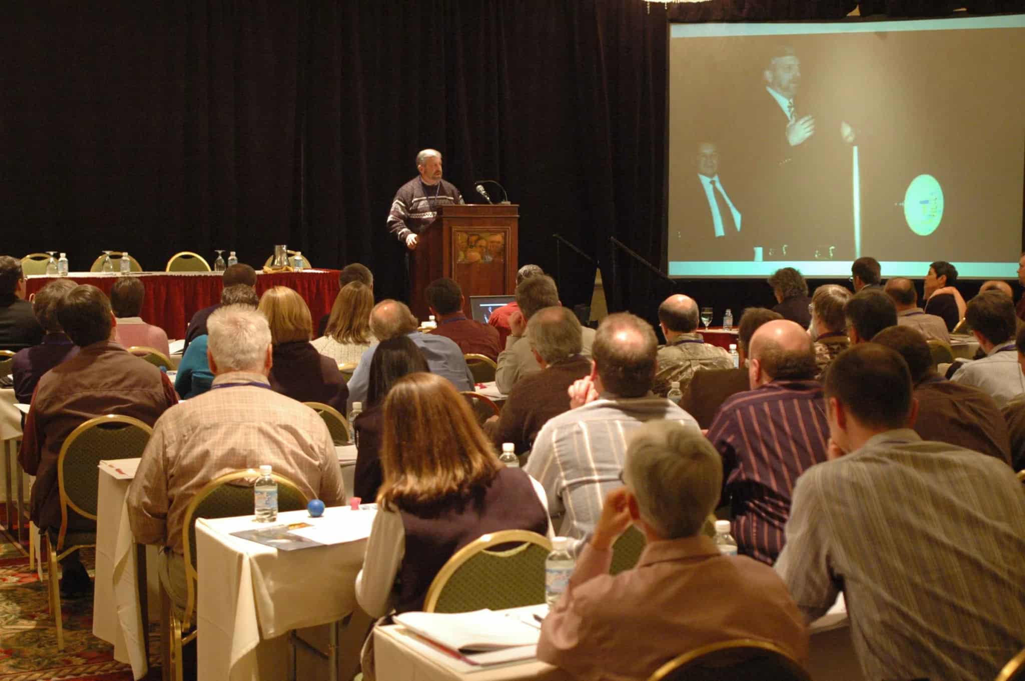 A man speaks at a podium to a large seated audience in a conference room.