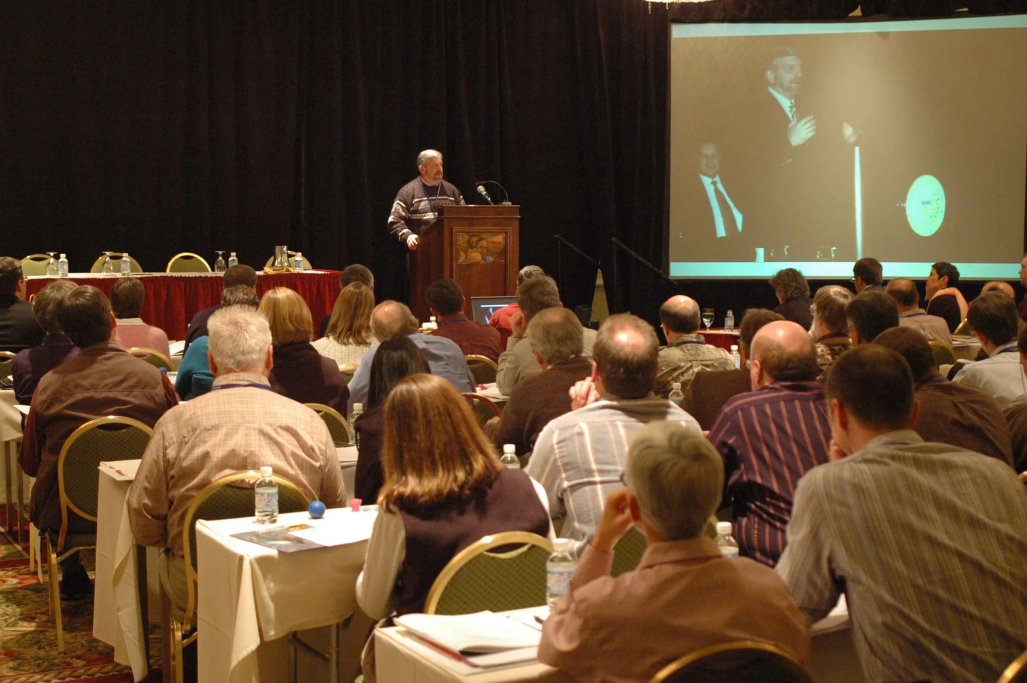 A man speaks at a podium to a large seated audience in a conference room.