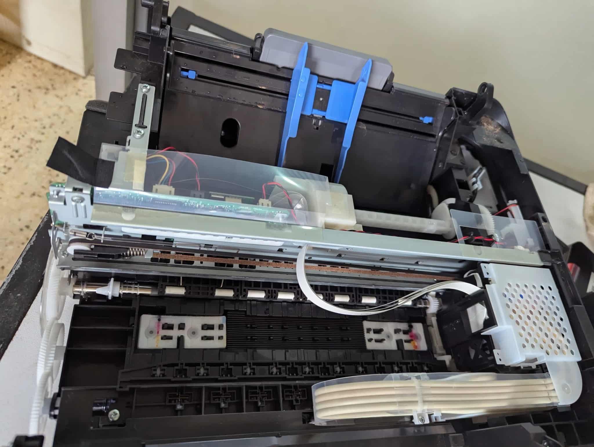 Close-up of the open internal mechanism of a printer or copier, showing gears, rollers, ribbon cables, and circuit boards.