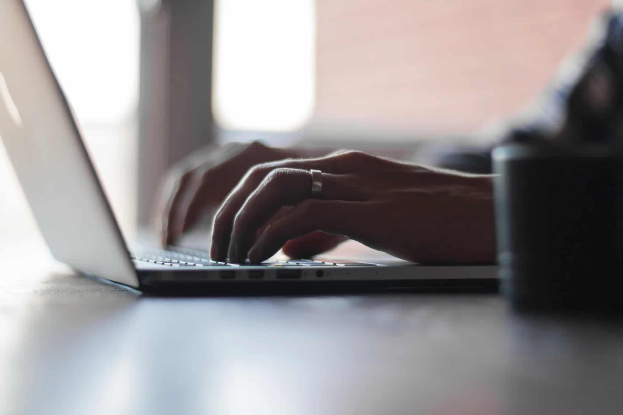 Close-up of two hands typing on a laptop keyboard at a low angle, with a mug blurred in the background.