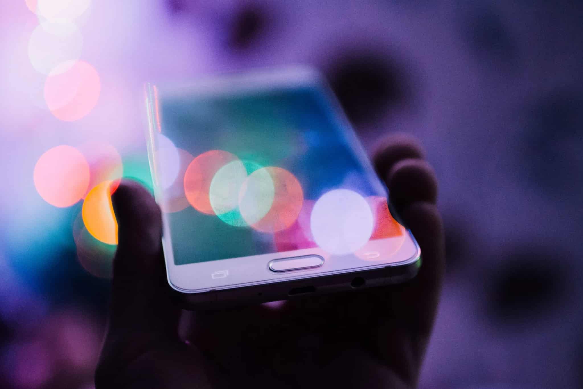 A hand holding a smartphone with a physical home button, photographed against a background of colorful blurred lights.