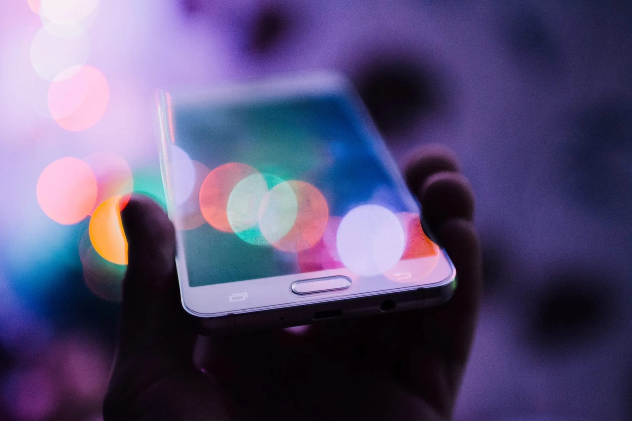 A hand holding a smartphone with a physical home button, photographed against a background of colorful blurred lights.
