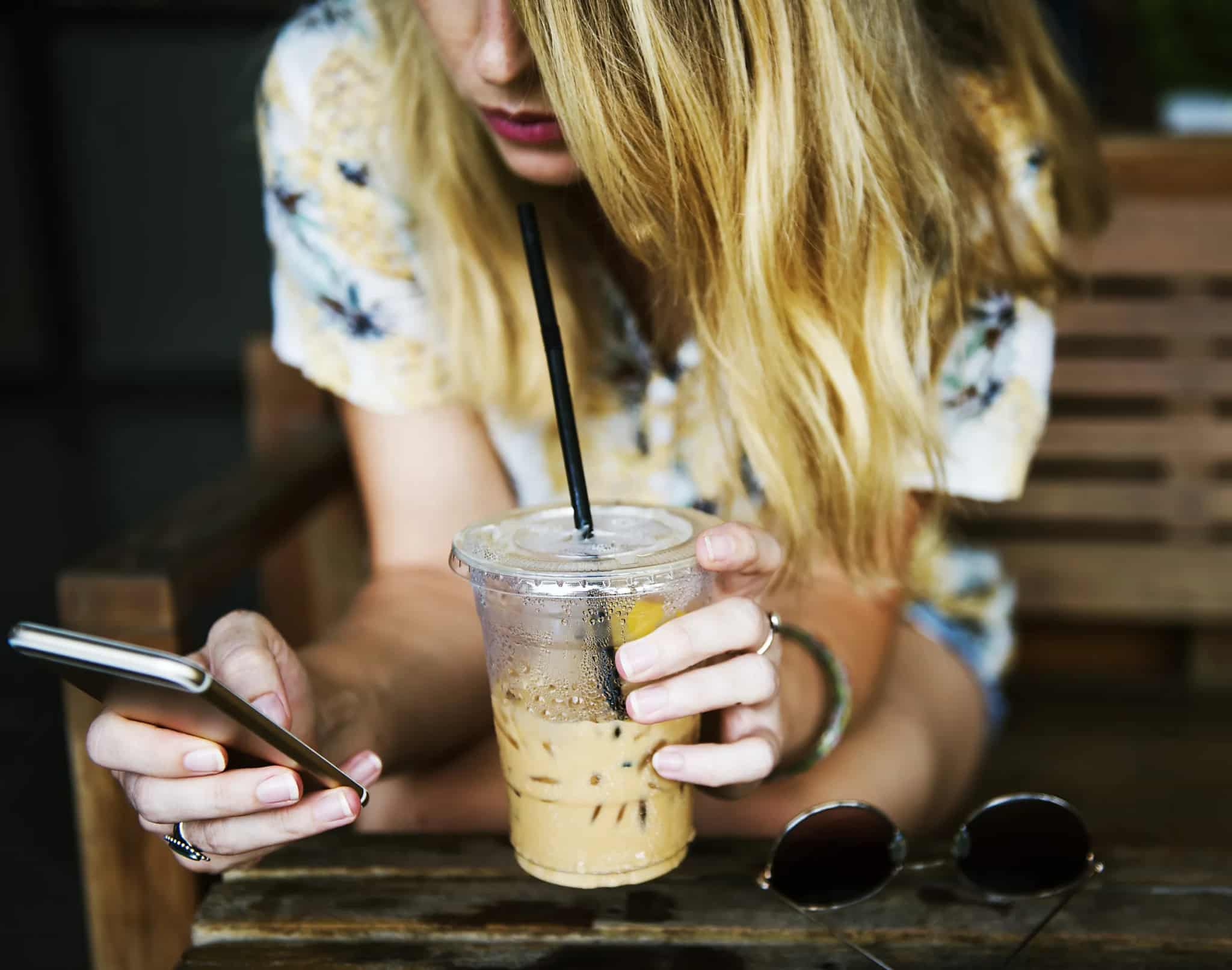 A person's hands holding a smartphone and an iced coffee at an outdoor table, with sunglasses resting nearby.