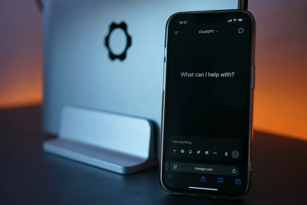A smartphone with an AI chat prompt on screen, propped beside a closed laptop on a desk.