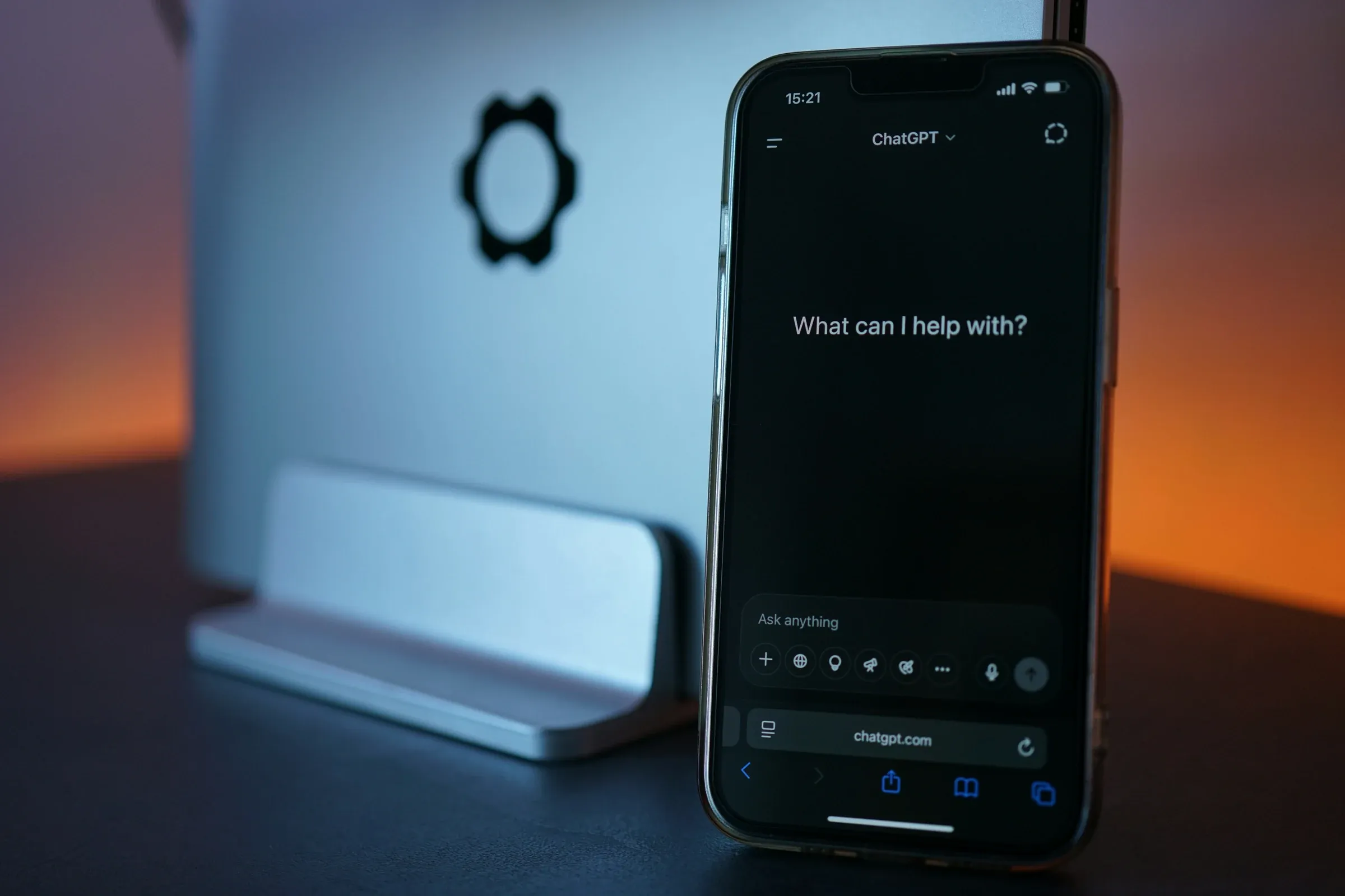 A smartphone with an AI chat prompt on screen, propped beside a closed laptop on a desk.