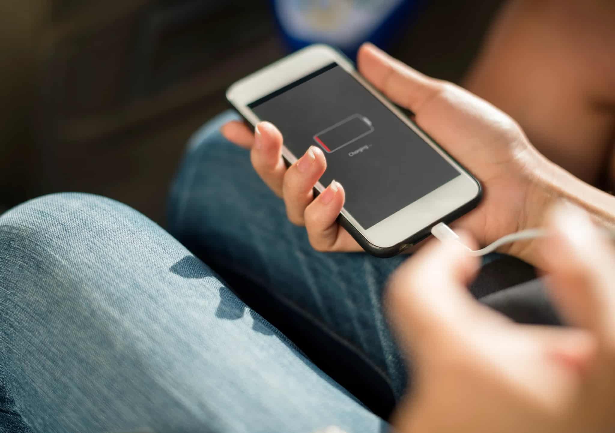 Close-up of hands holding a smartphone displaying a low battery charging icon, with a charging cable plugged in.