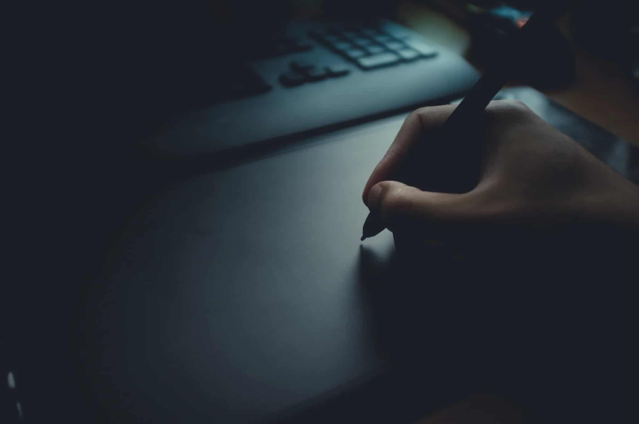 A hand holding a stylus poised over a dark graphics tablet, with a keyboard visible in the blurred background.