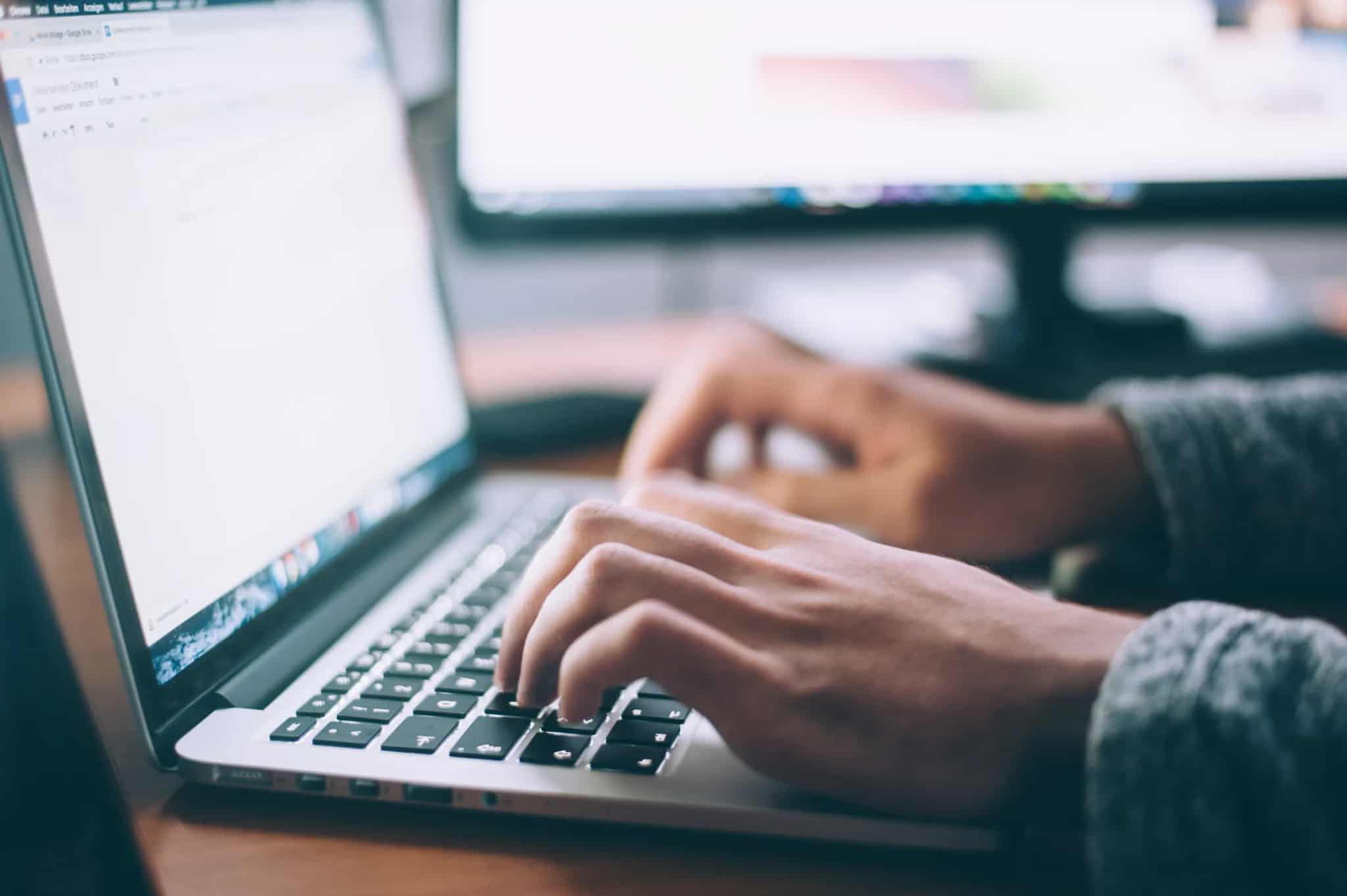 Close-up of hands typing on a laptop keyboard with a second monitor glowing in the background.