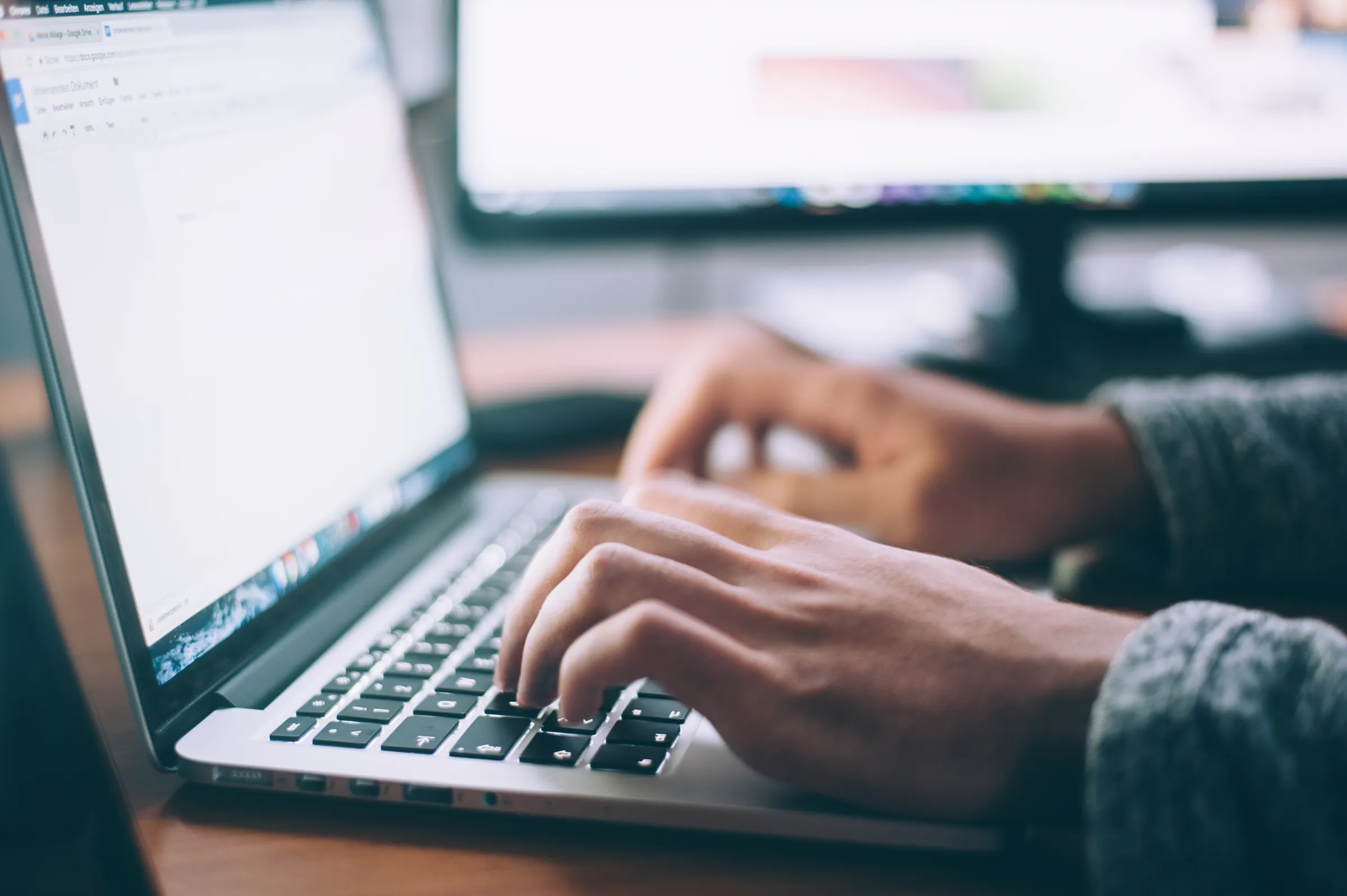 Close-up of hands typing on a laptop keyboard with a second monitor glowing in the background.