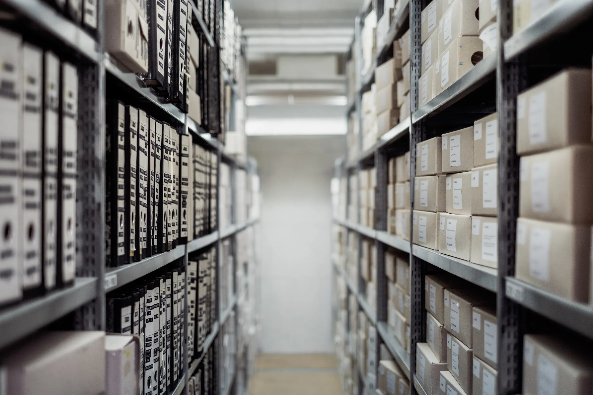 A narrow aisle between metal shelving units filled with labeled archive boxes and binders.