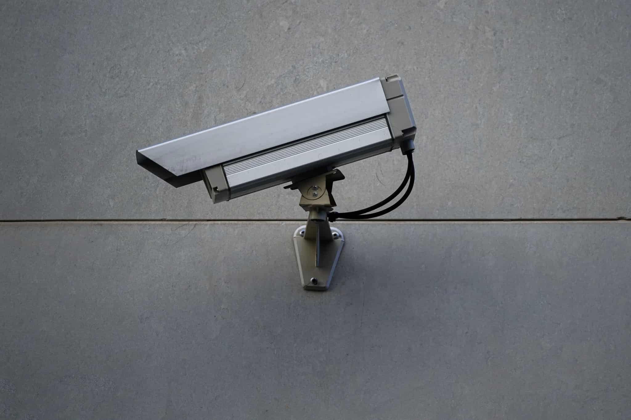 A silver CCTV security camera mounted on a bracket against a plain gray stone wall.