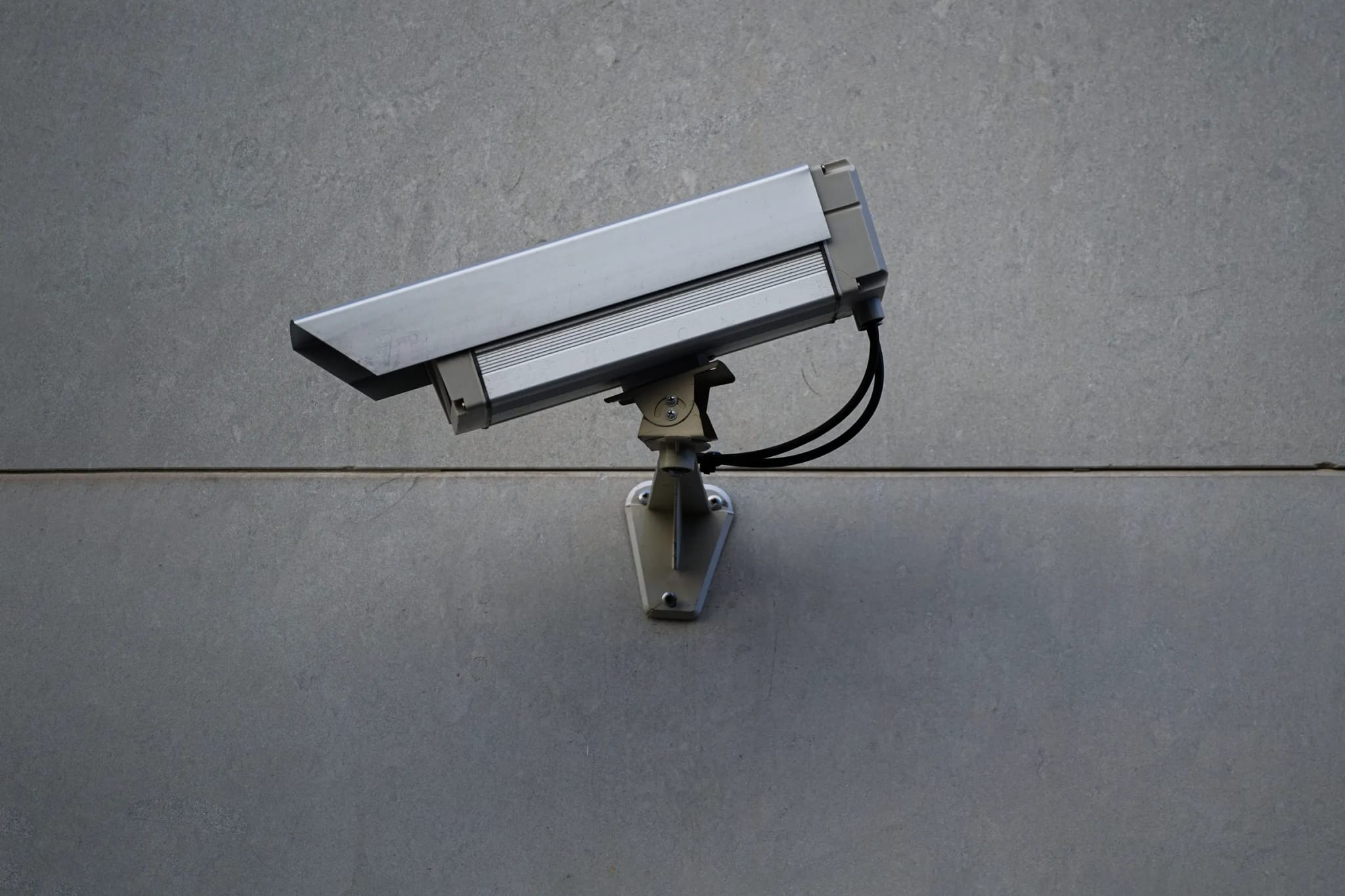 A silver CCTV security camera mounted on a bracket against a plain gray stone wall.