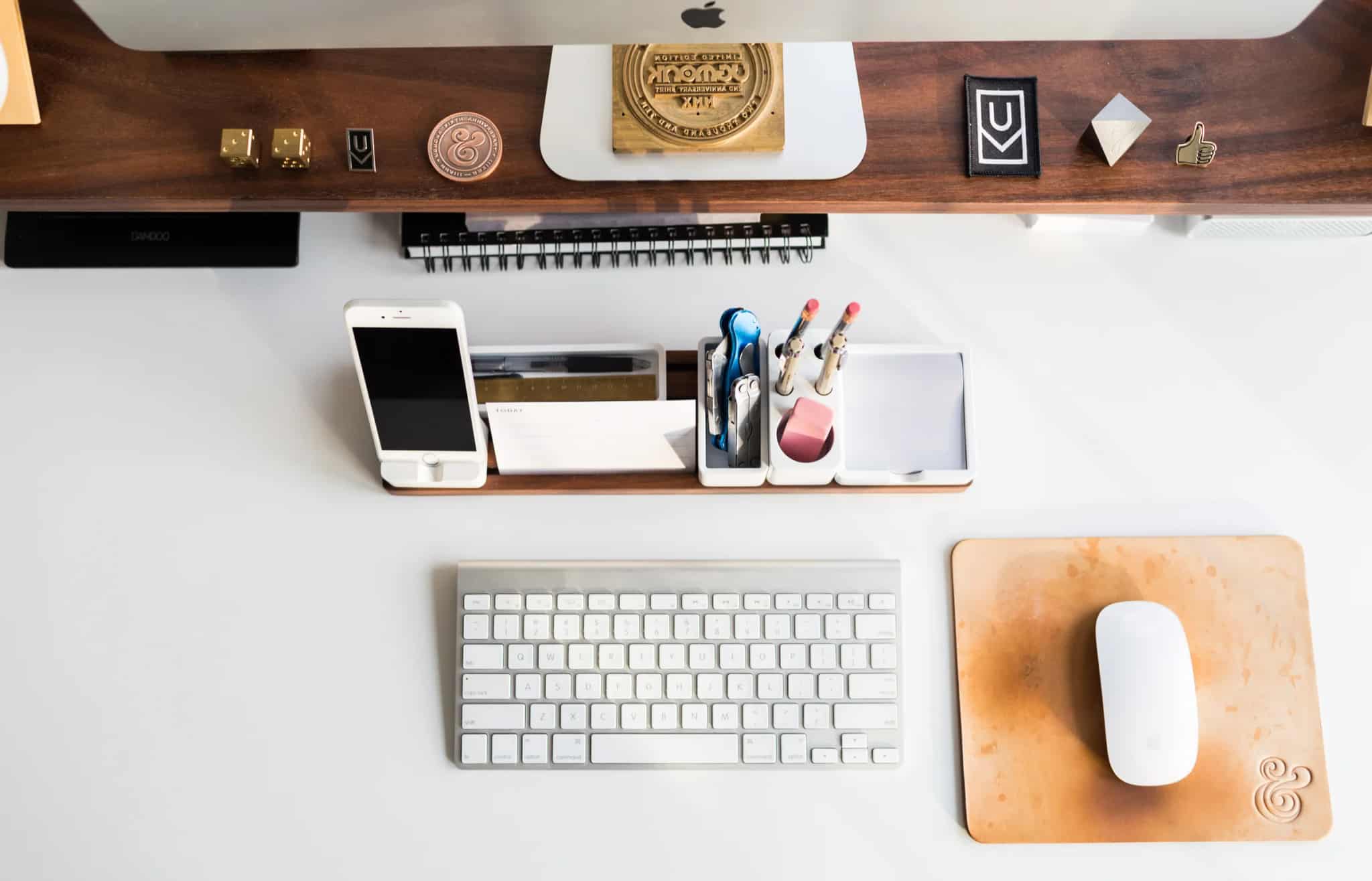 An overhead view of an iMac, keyboard, mouse and phone dock on a tidy desk, with small trinkets lined up on a wooden shelf above.