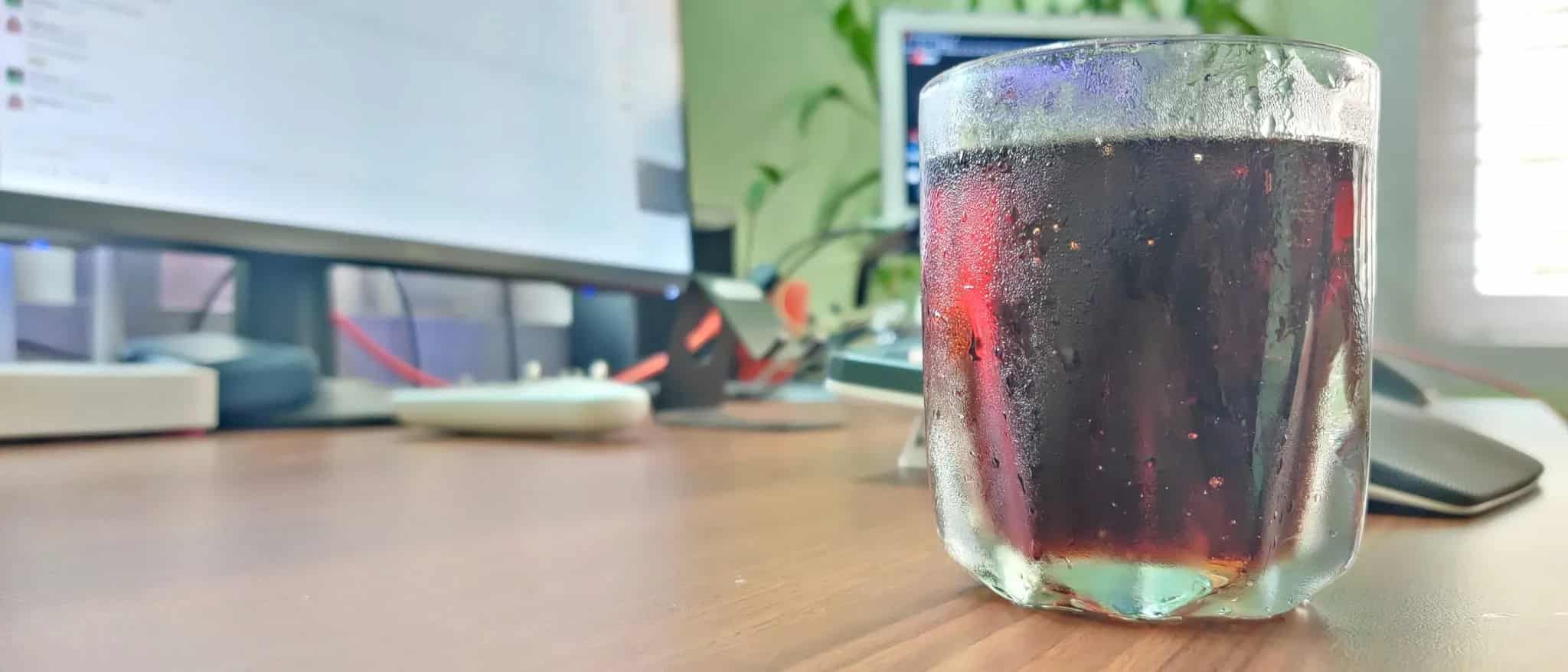 A glass of iced dark soda sits on a wooden desk in front of blurred computer monitors and green houseplants.