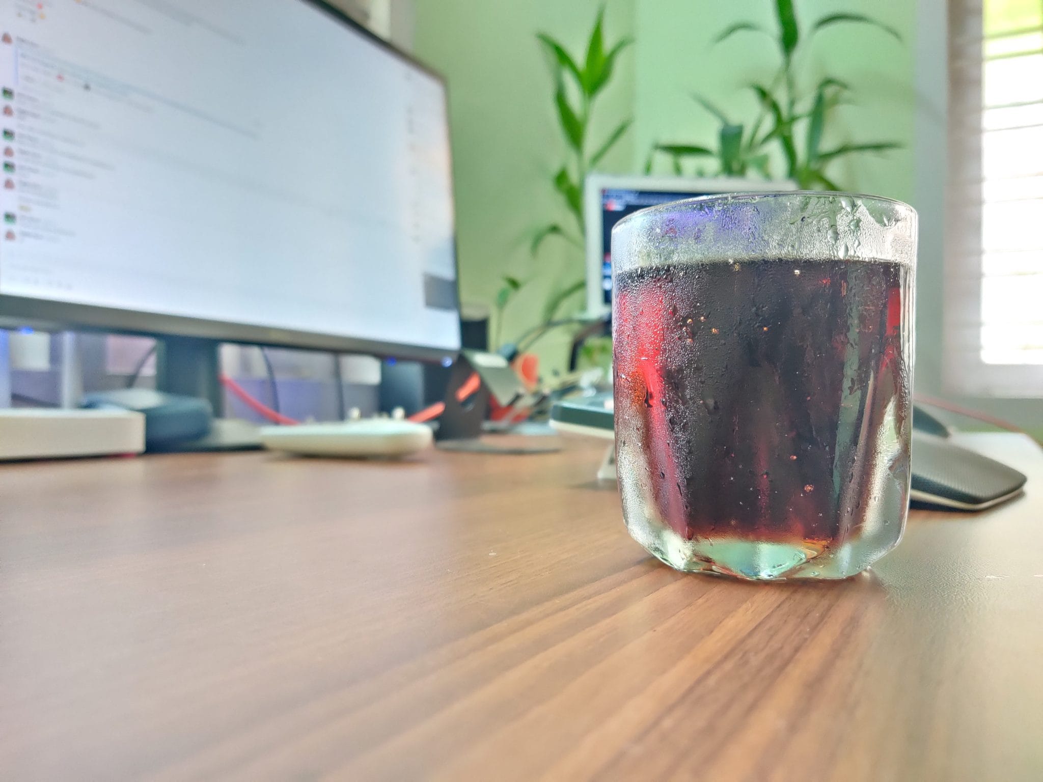 A glass of iced dark soda sits on a wooden desk in front of blurred computer monitors and green houseplants.