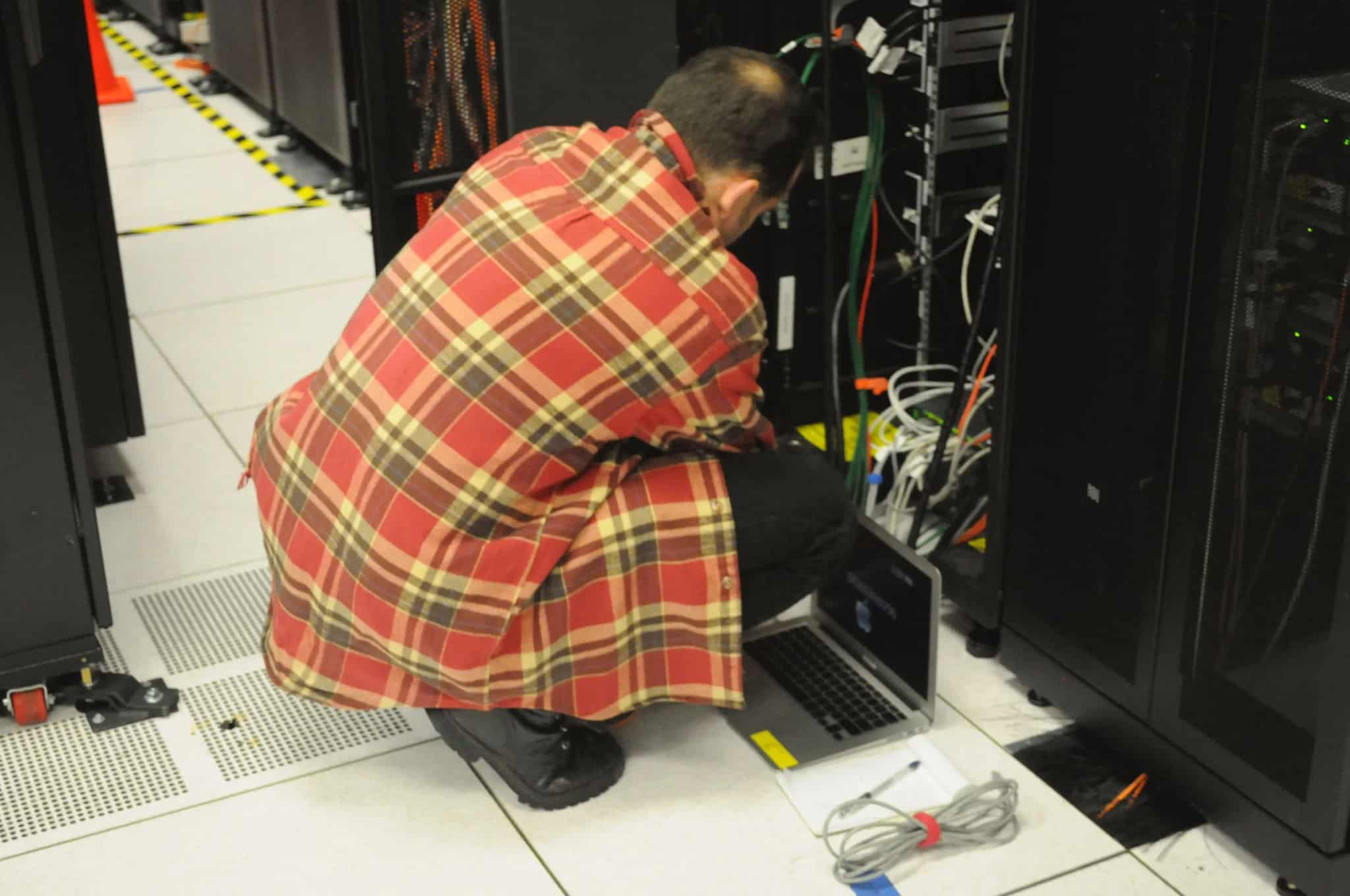 An IT technician crouches beside an open server rack, checking equipment with a laptop on the floor beside him.