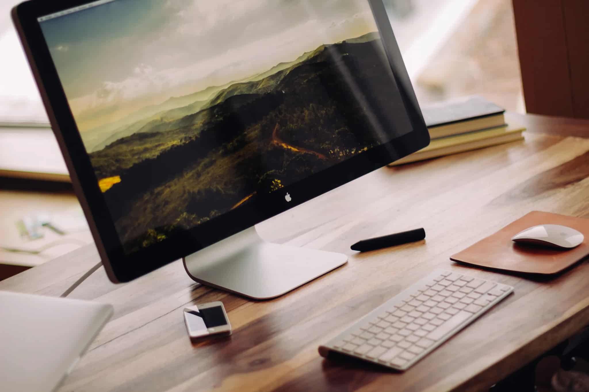 An angled desktop monitor displaying a hazy mountain-forest wallpaper, next to a wireless keyboard, mouse, stylus and smartphone on a wooden desk.