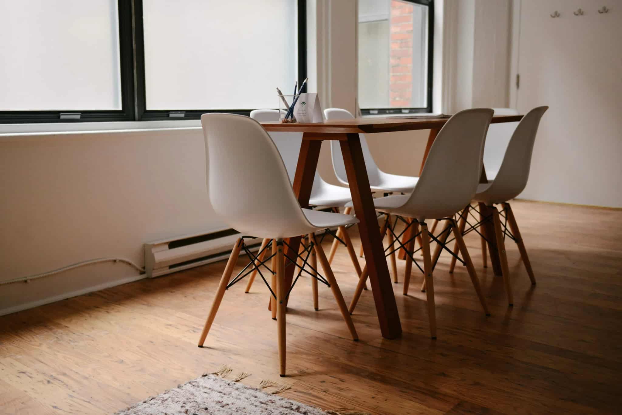 A bright, modern meeting room with a wooden table surrounded by white chairs, pencils in a cup and a WiFi-password card on the table, lit by tall windows.