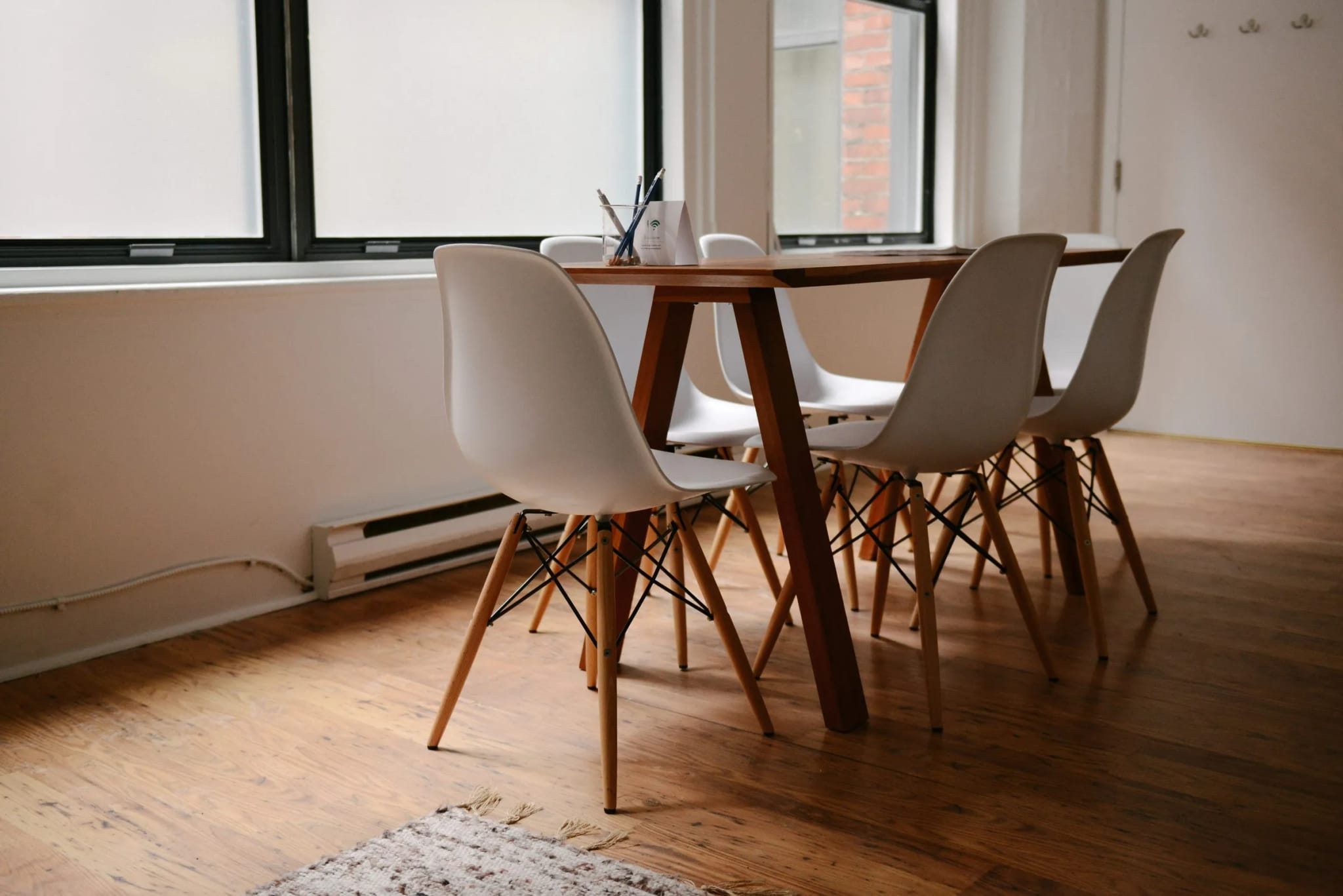 A bright, modern meeting room with a wooden table surrounded by white chairs, pencils in a cup and a WiFi-password card on the table, lit by tall windows.