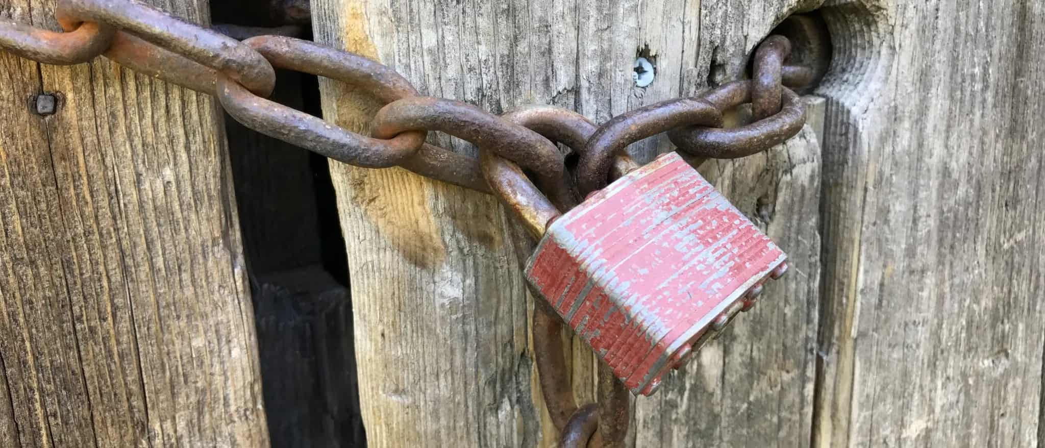A weathered red padlock secures a heavy rusted chain wrapped around a wooden gate post.