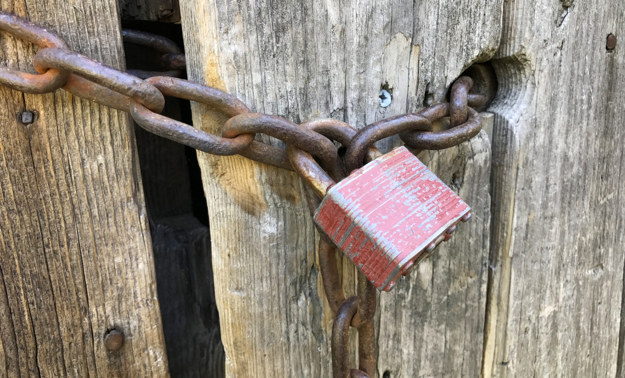 A weathered red padlock secures a heavy rusted chain wrapped around a wooden gate post.