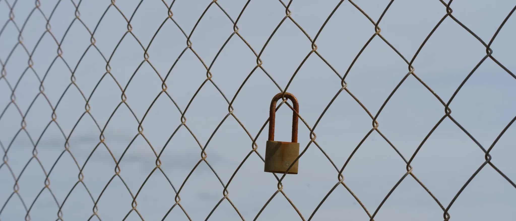 A small rusty padlock hanging on a chain-link fence against the sky.
