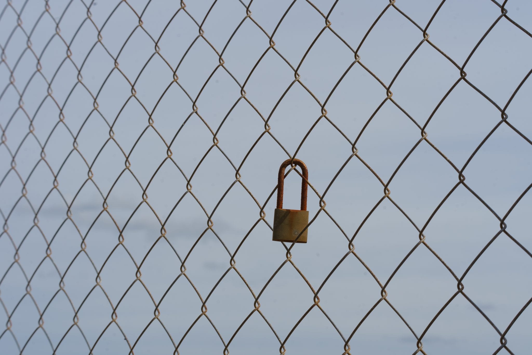 A small rusty padlock hanging on a chain-link fence against the sky.