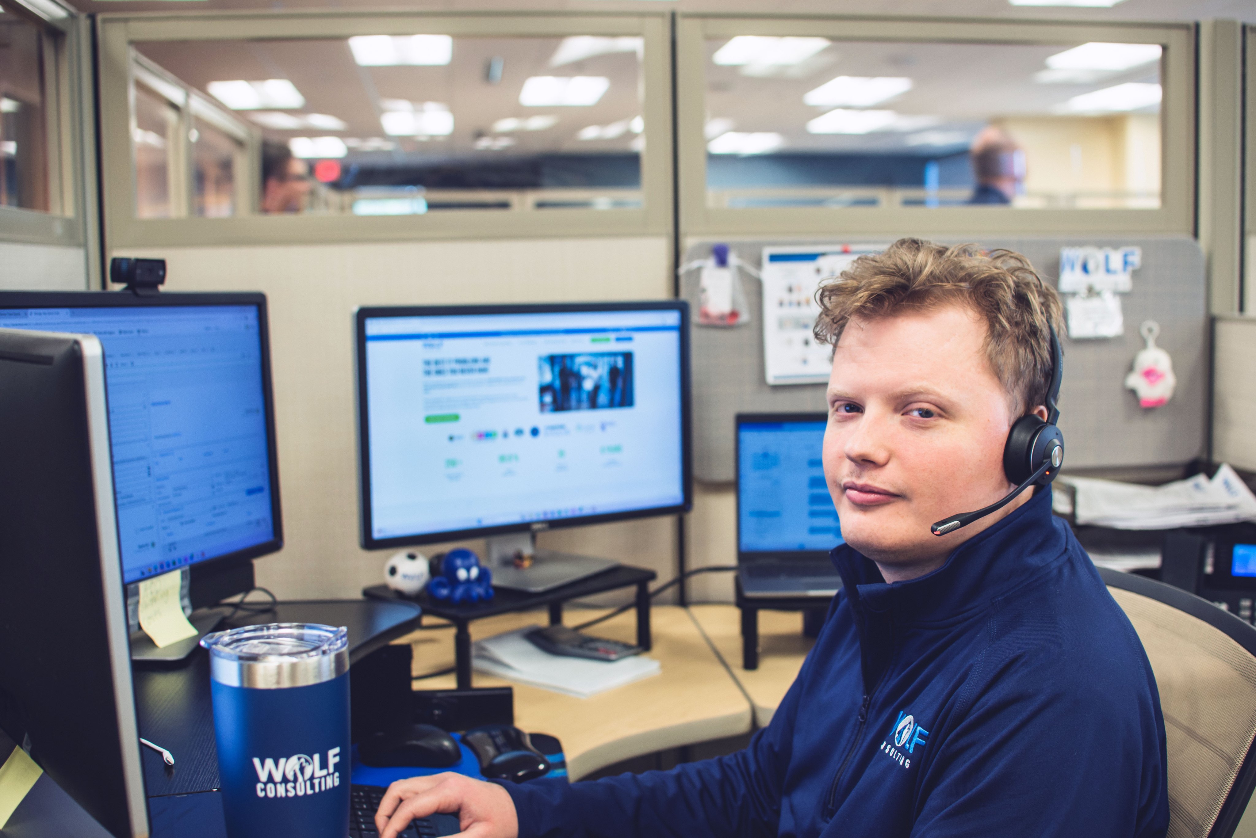 A Wolf Consulting support engineer wearing a headset at a multi-monitor desk, a WOLF Consulting tumbler in the foreground